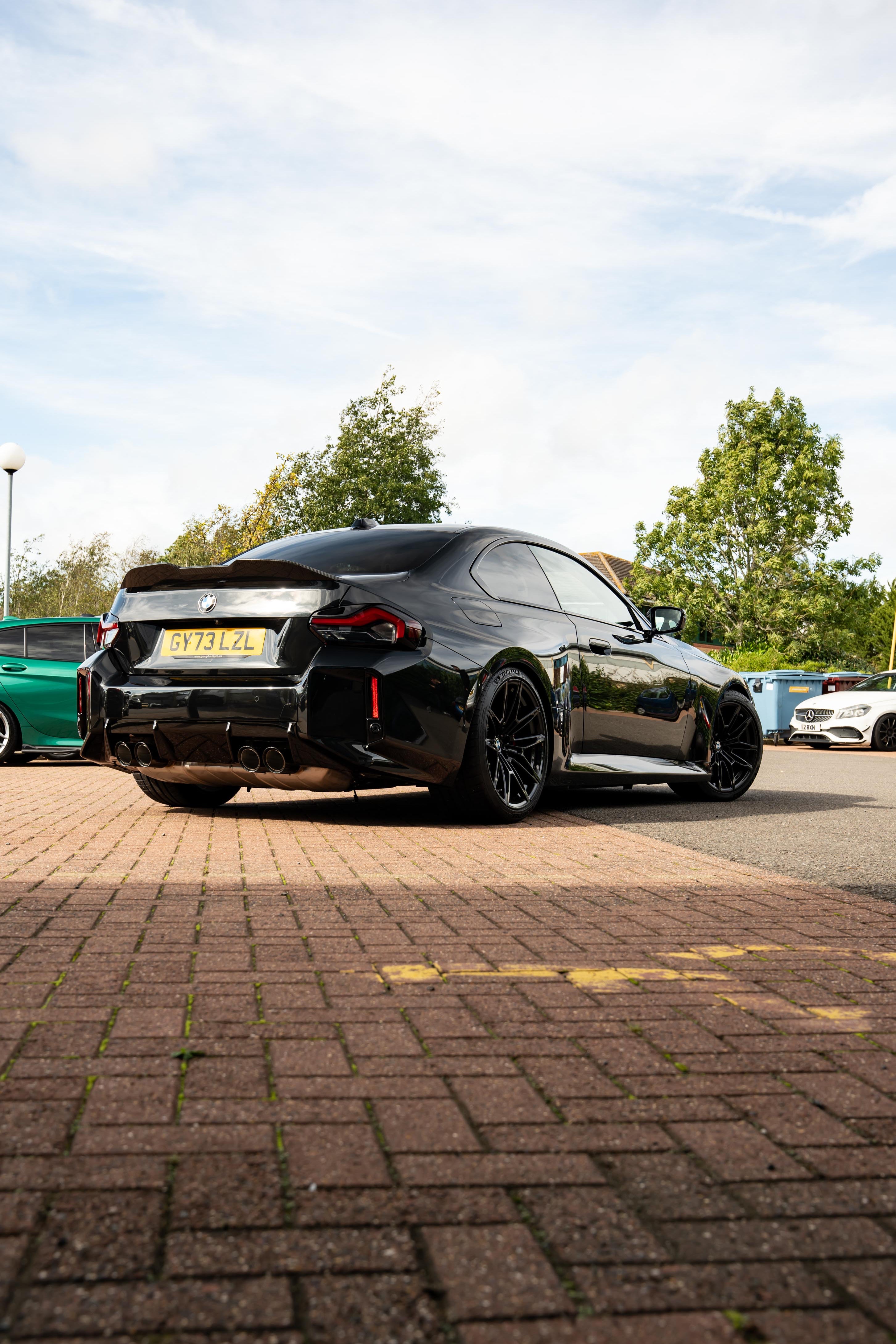 Black BMW M2 G87 parked on a paved area with trees and other cars in the background