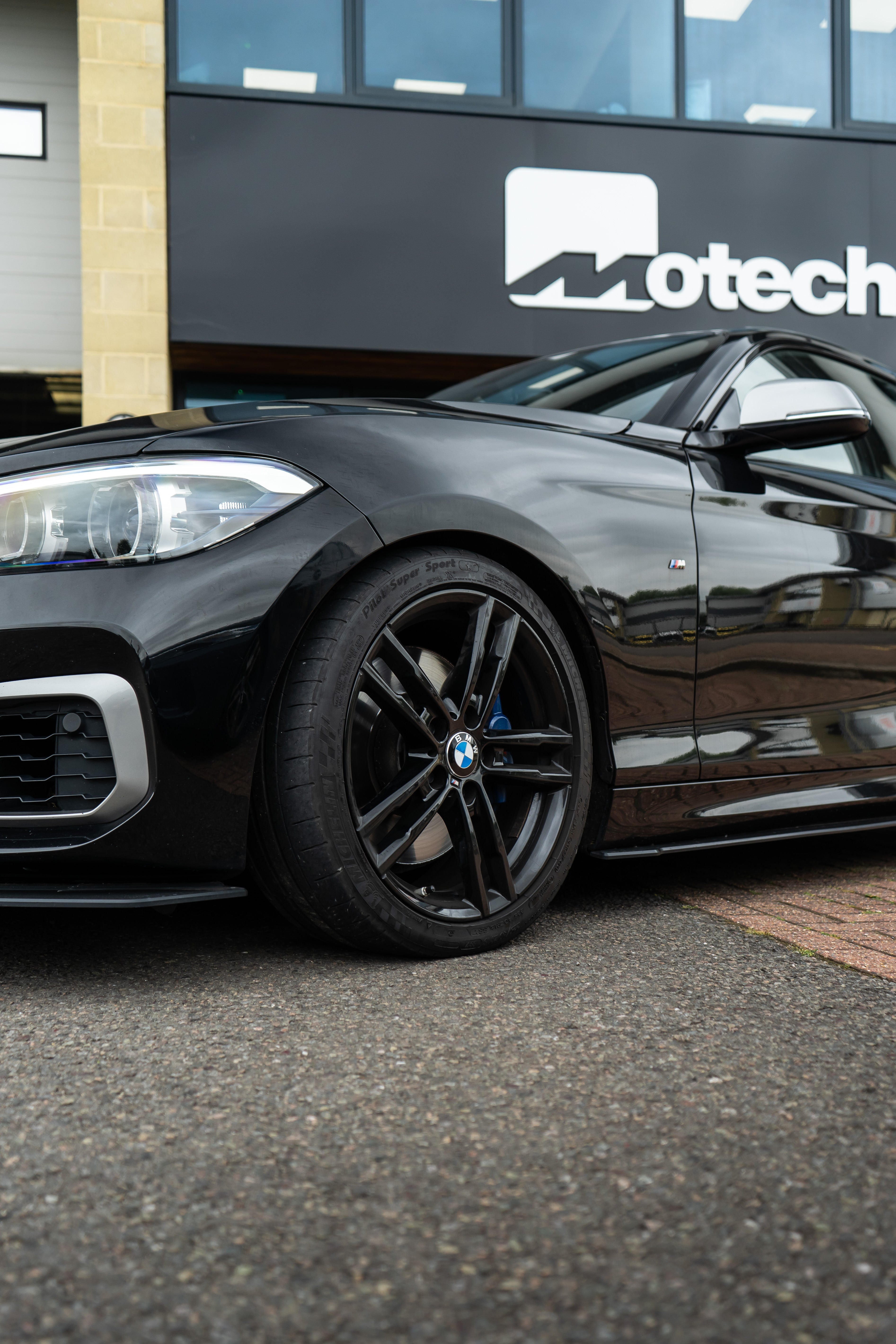 A close-up shot of a black BMW's wheel outside a building with a sign that says 'Motech'.