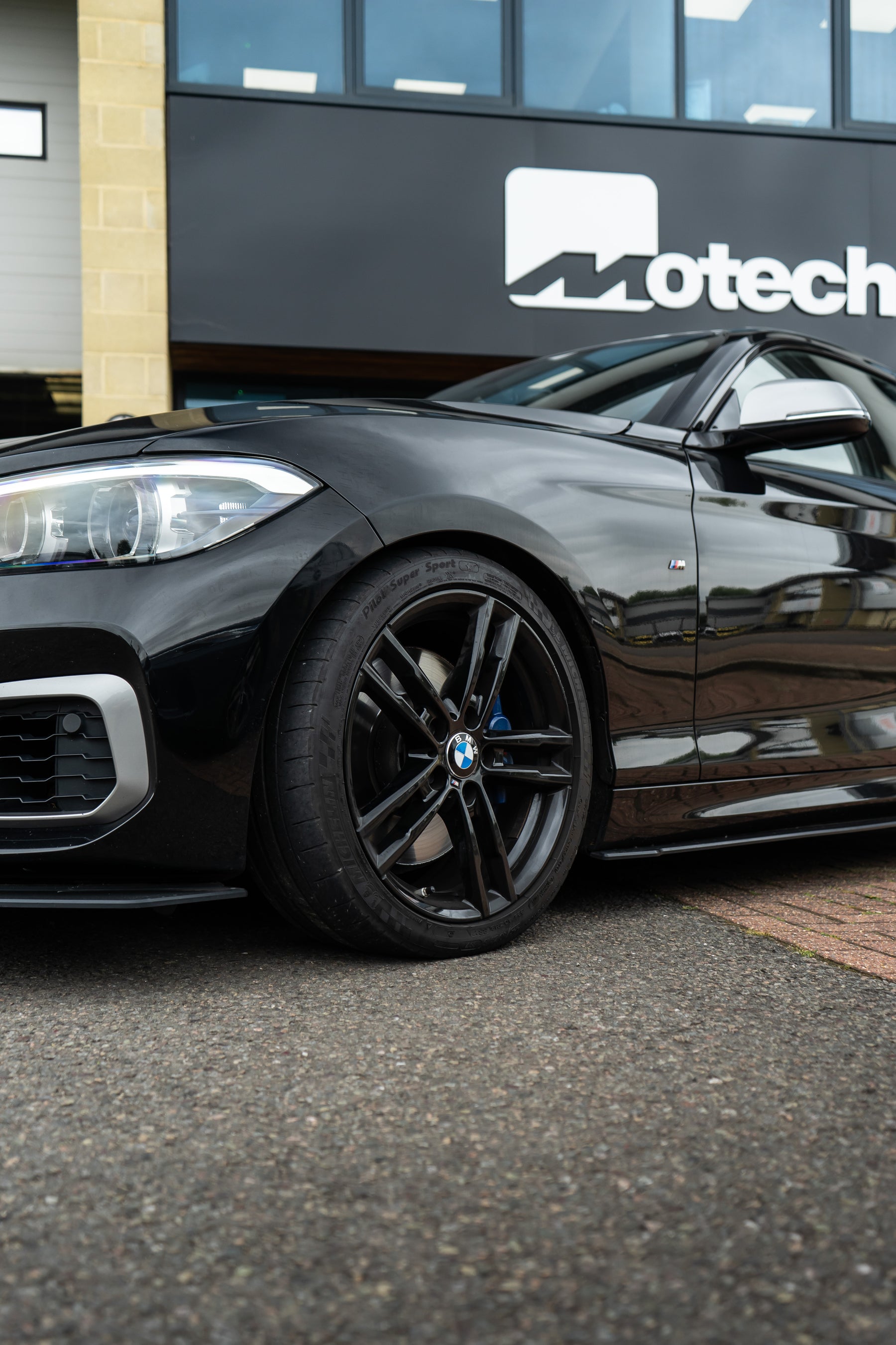 A close-up shot of a black BMW's wheel outside a building with a sign that says 'Motech'.