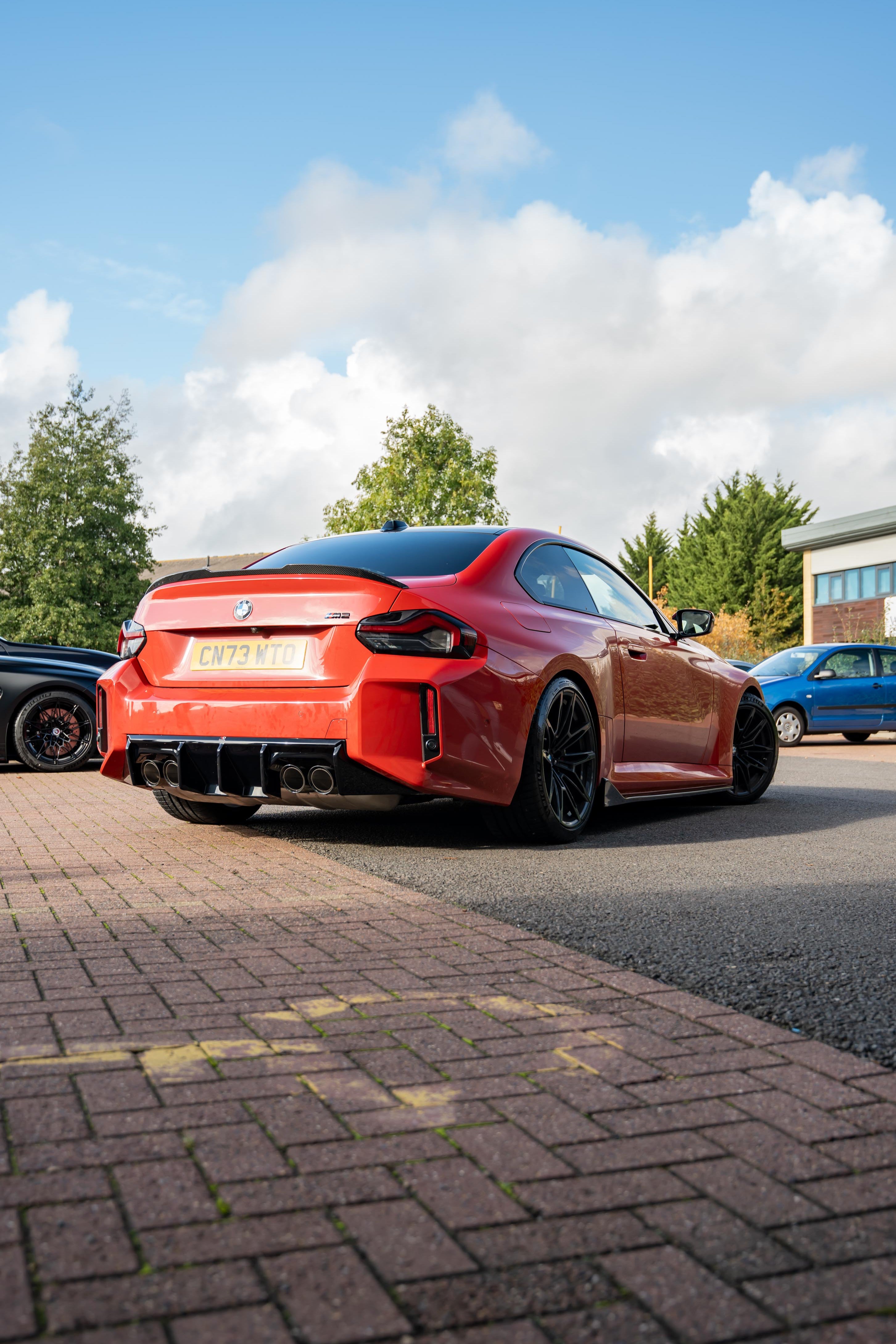 Red BMW M2 G87 with Motech stance springs parked on a brick driveway with a blue sky and trees in the background.
