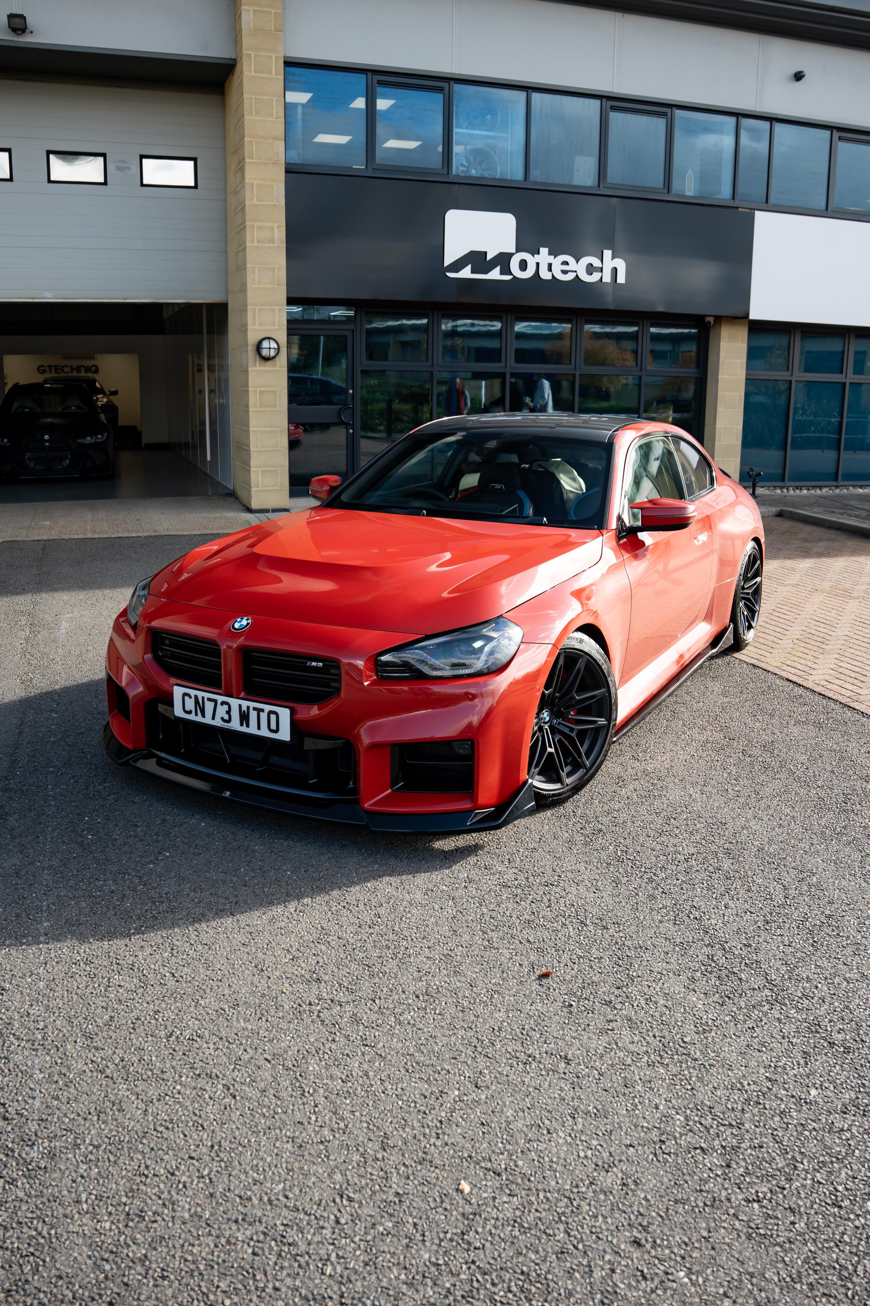 Red BMW M2 G87 on Motech Stance Springs parked outside a building with 'Motech' branding.
