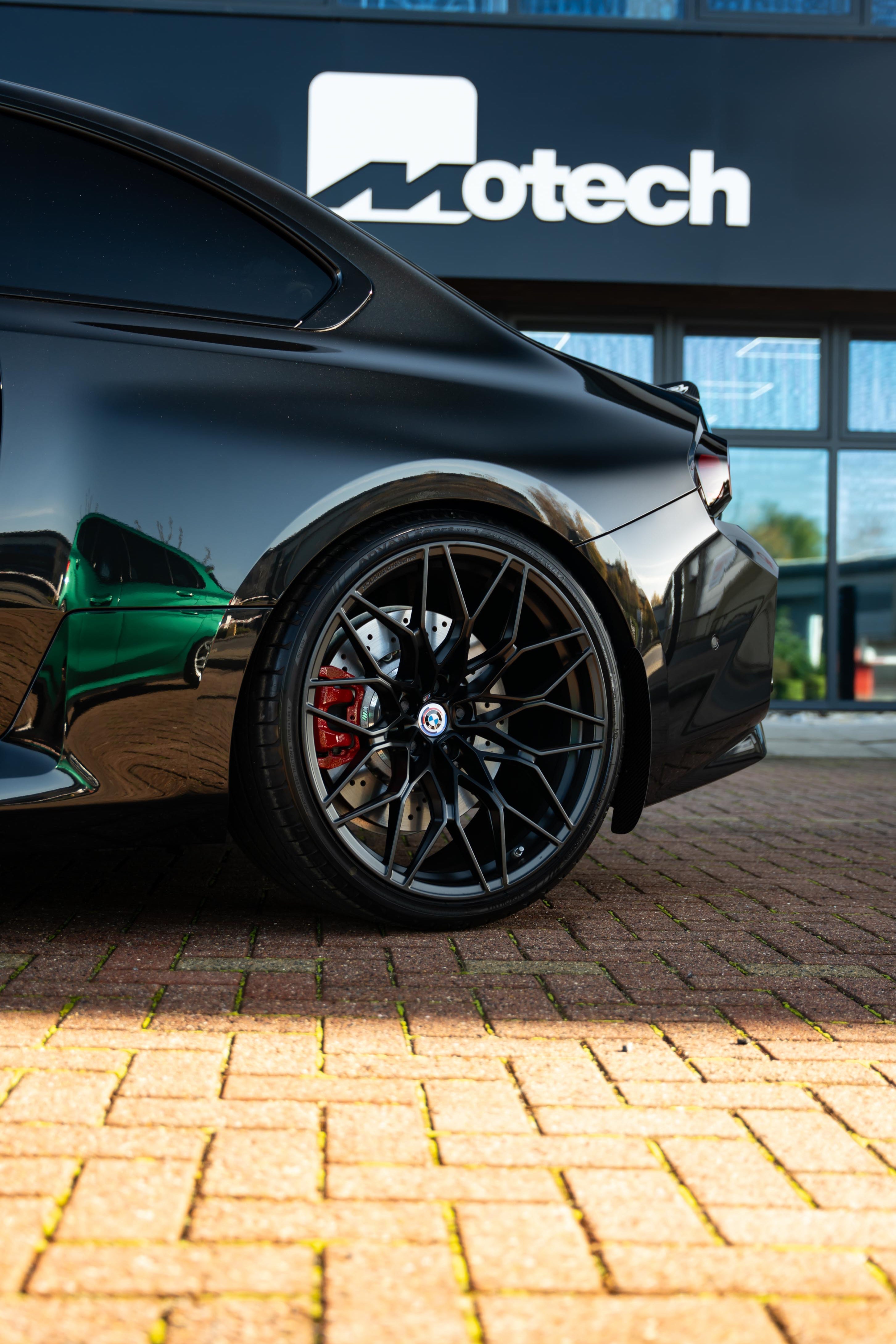 Car wheel on a G87 M2 with black rim and red accents on a brick surface, Motech logo in the background.