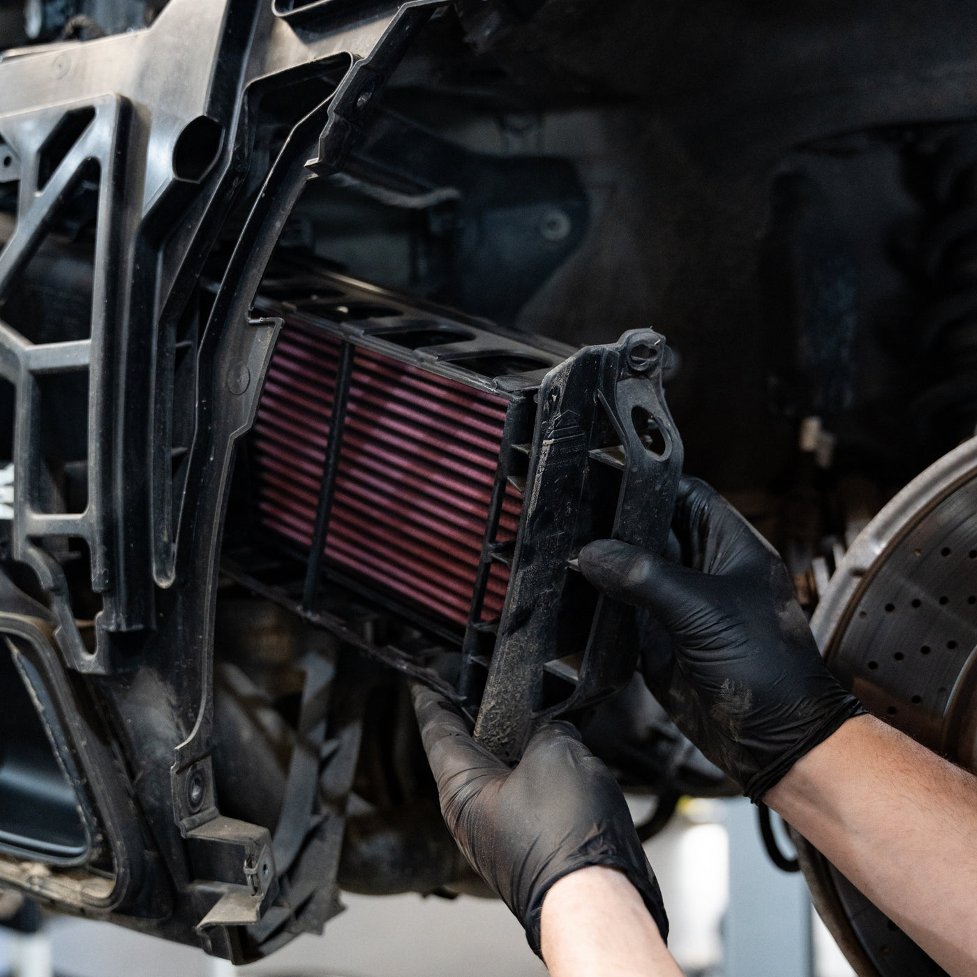 Person wearing black gloves installing an air filter into a vehicle's engine bay.