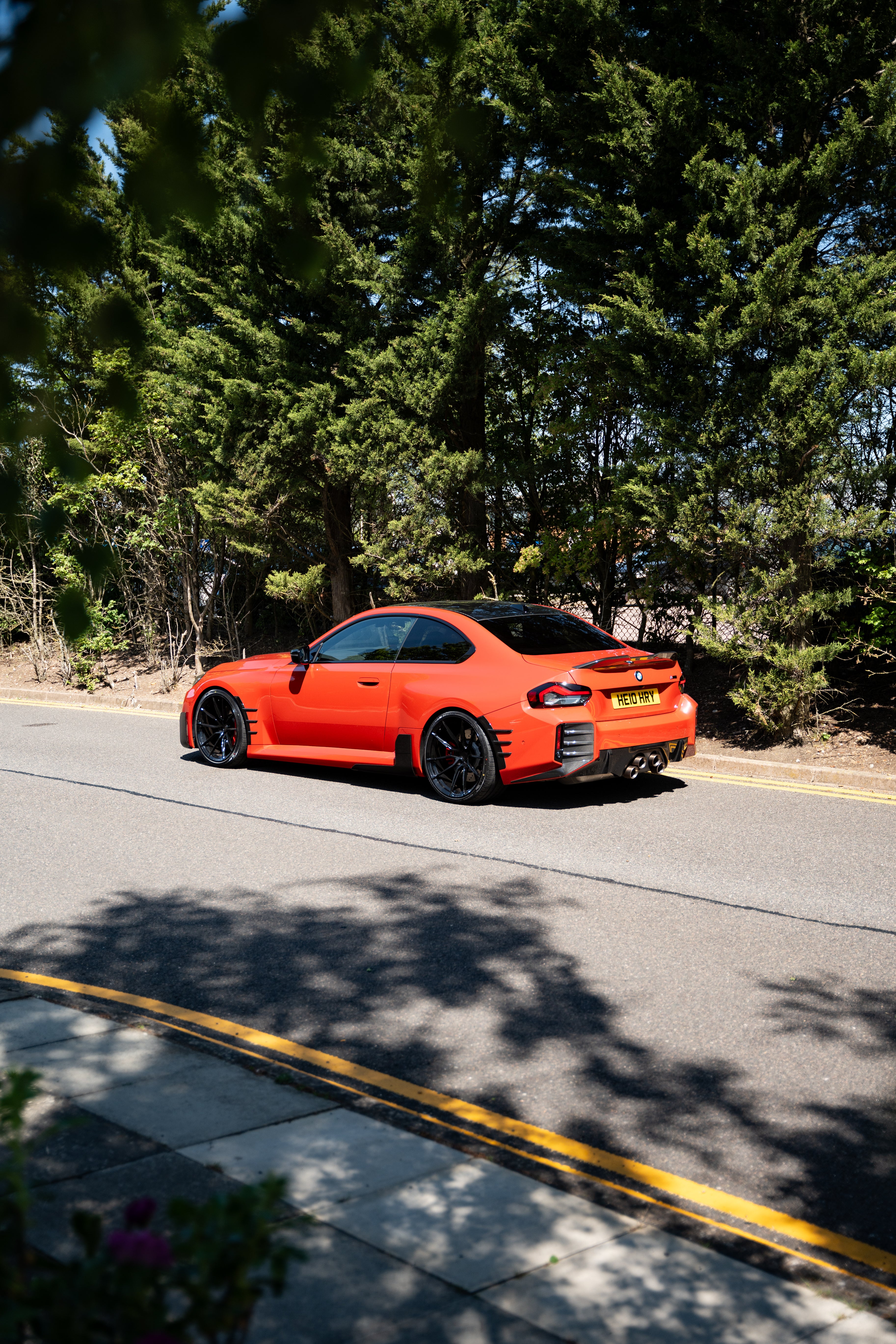 Orange BMW M2 G87 on Motech Stance Springs parked by trees and bushes.