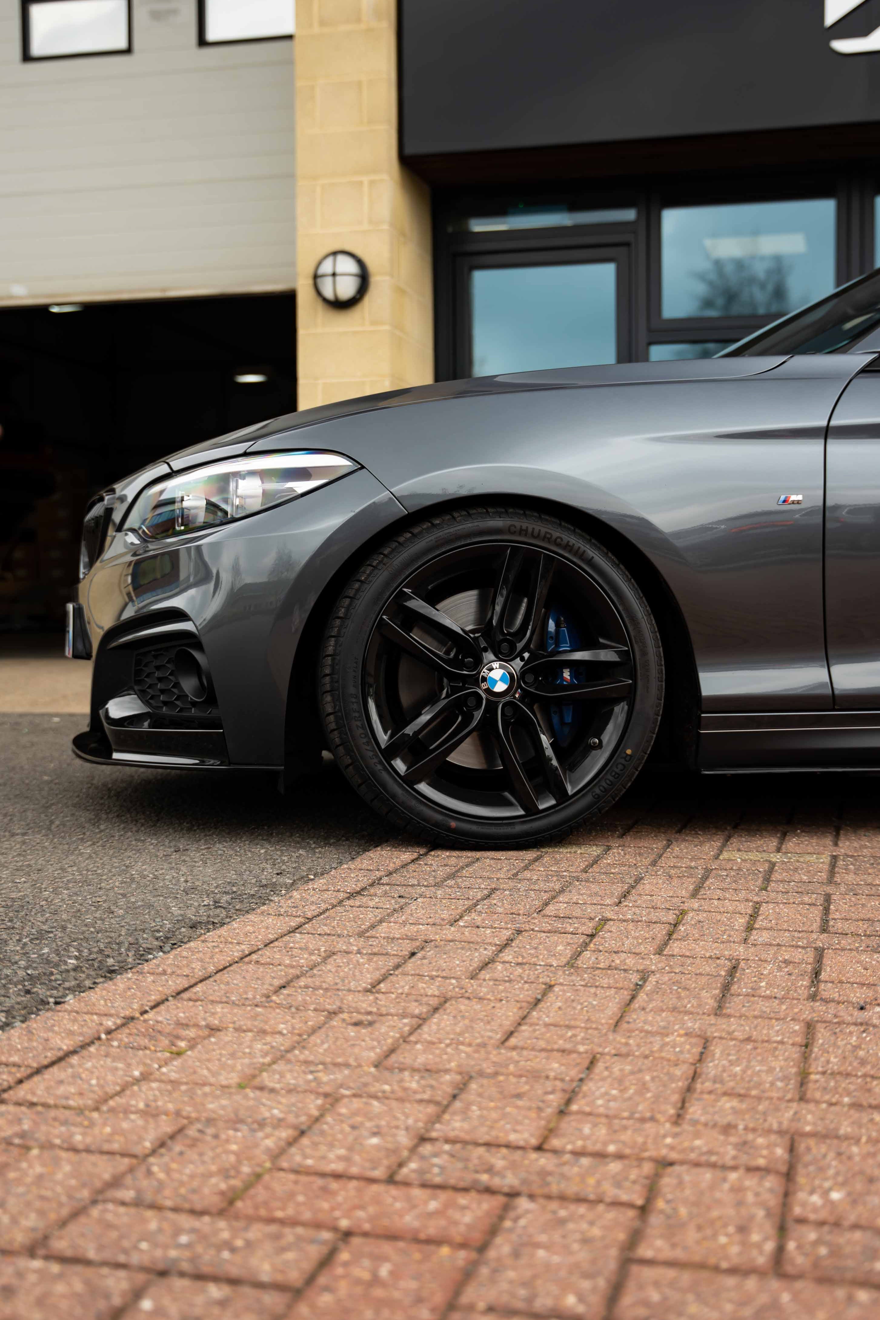 Grey BMW car on Motech Stance Springs with black rims parked on a brick pavement in front of a building.
