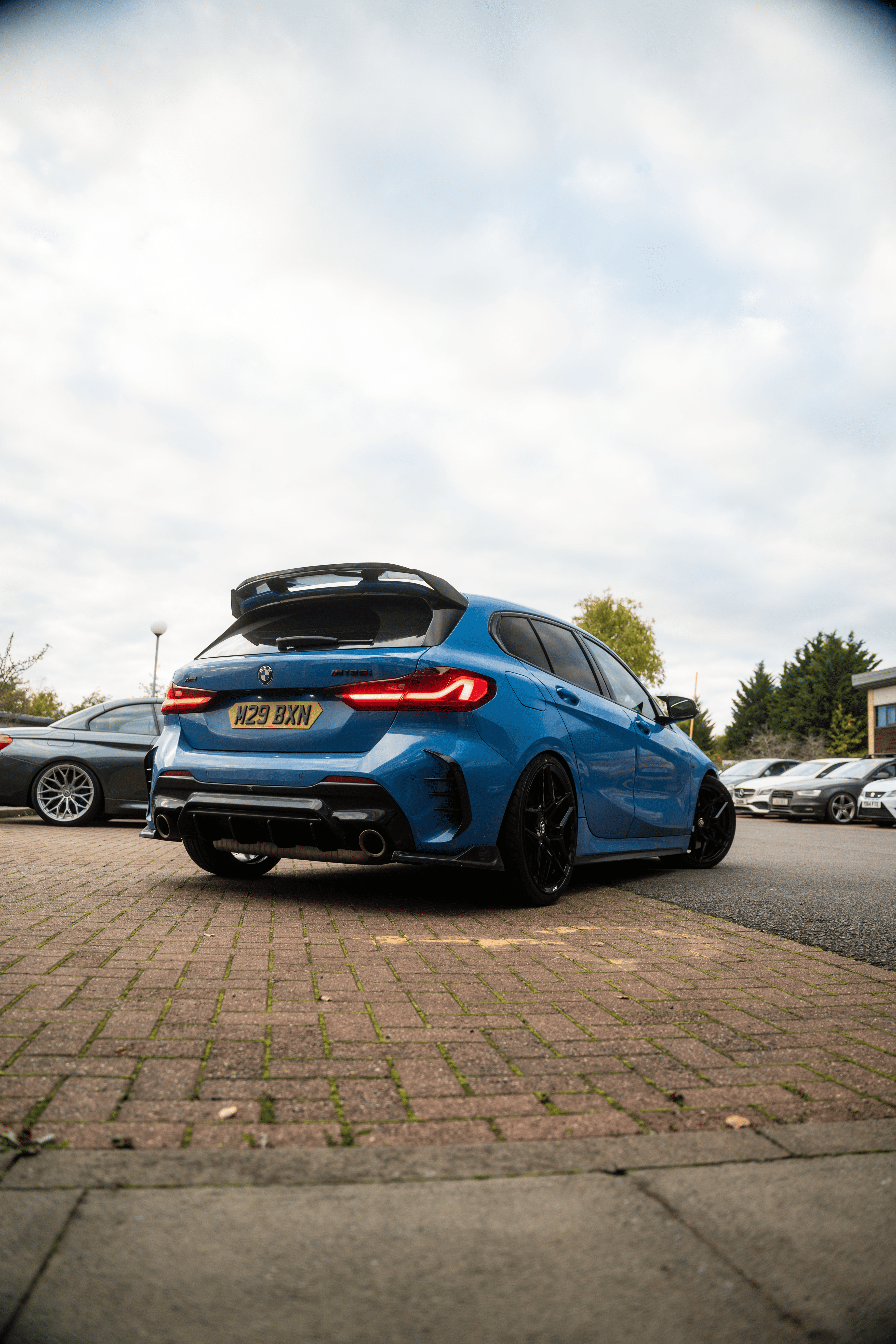 Blue BMW parked on a brick driveway with trees and other cars in the background
