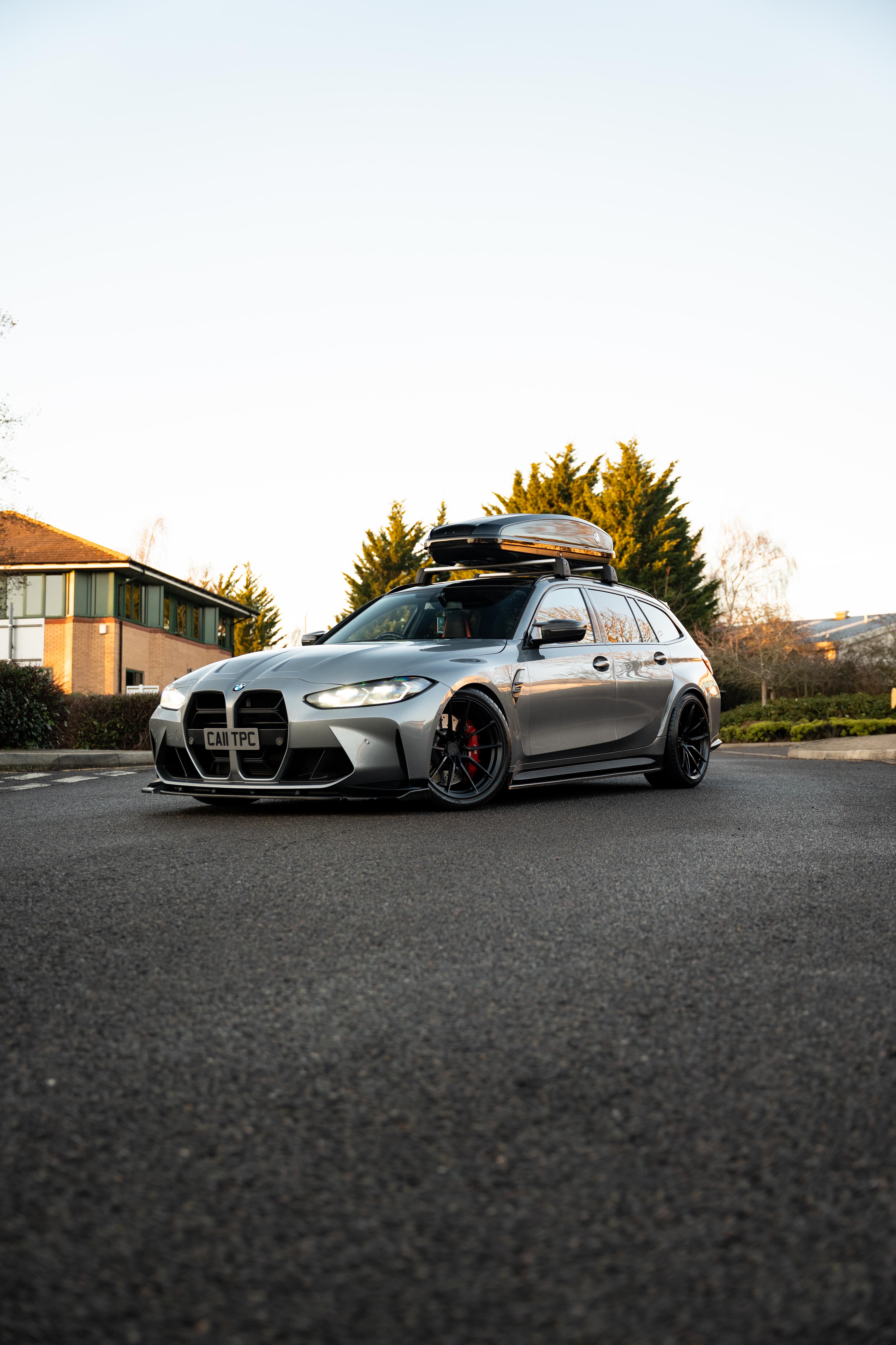 Front of a Grey G81 M3 Touring with a roof box & is sat on Motech stance springs - parked outside as the sun is setting on some trees.