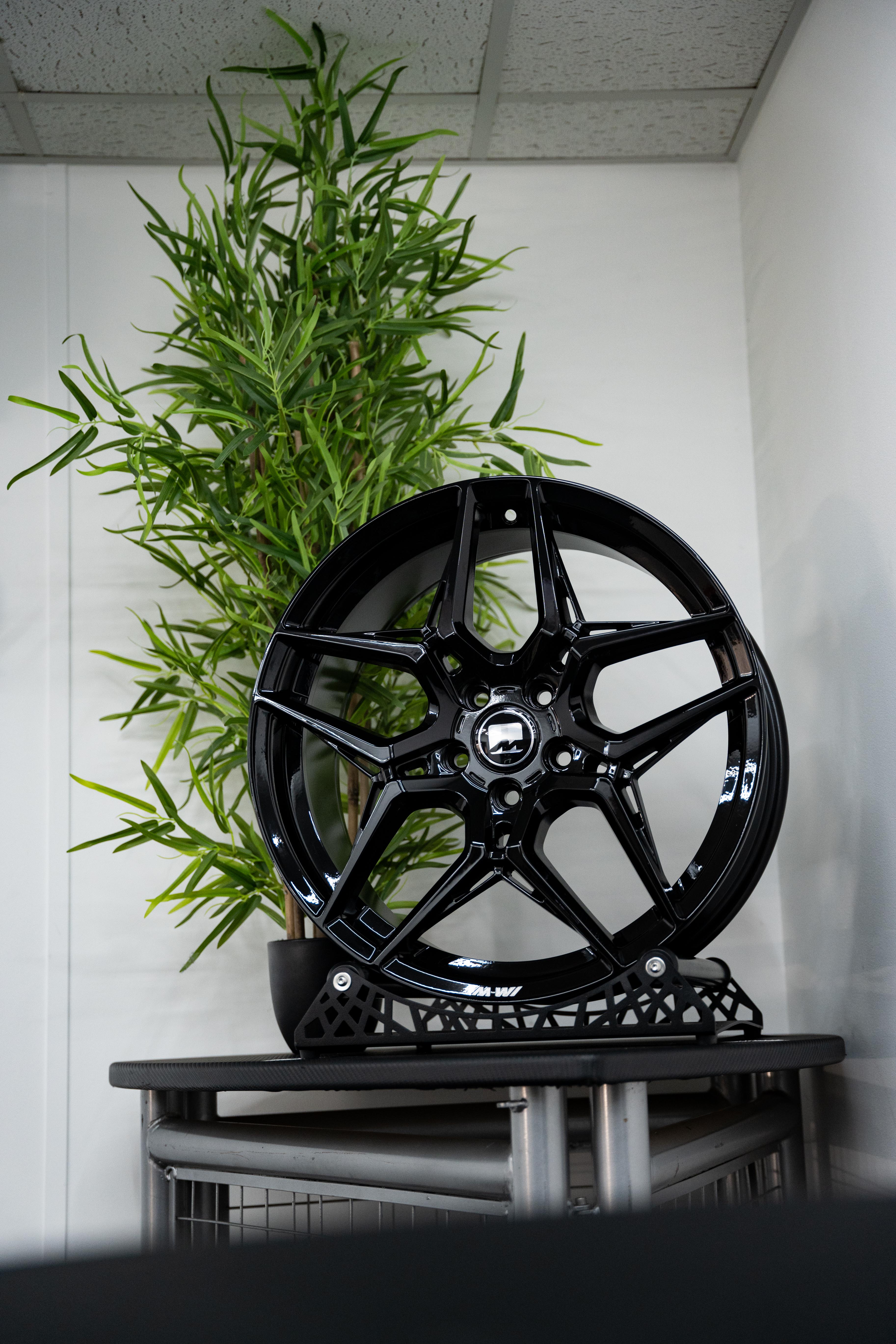 A single Precision Black M-W1 Wheel displayed on a stand with a green plant in the background.