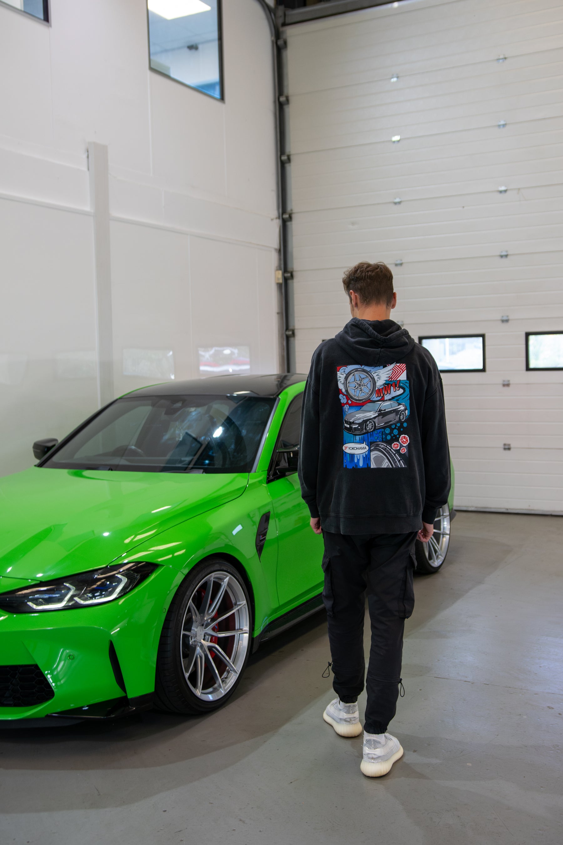 Person wearing a Stone-washed Treadlight collection Motech hoodie with a colorful design, standing next to a green car in a garage.
