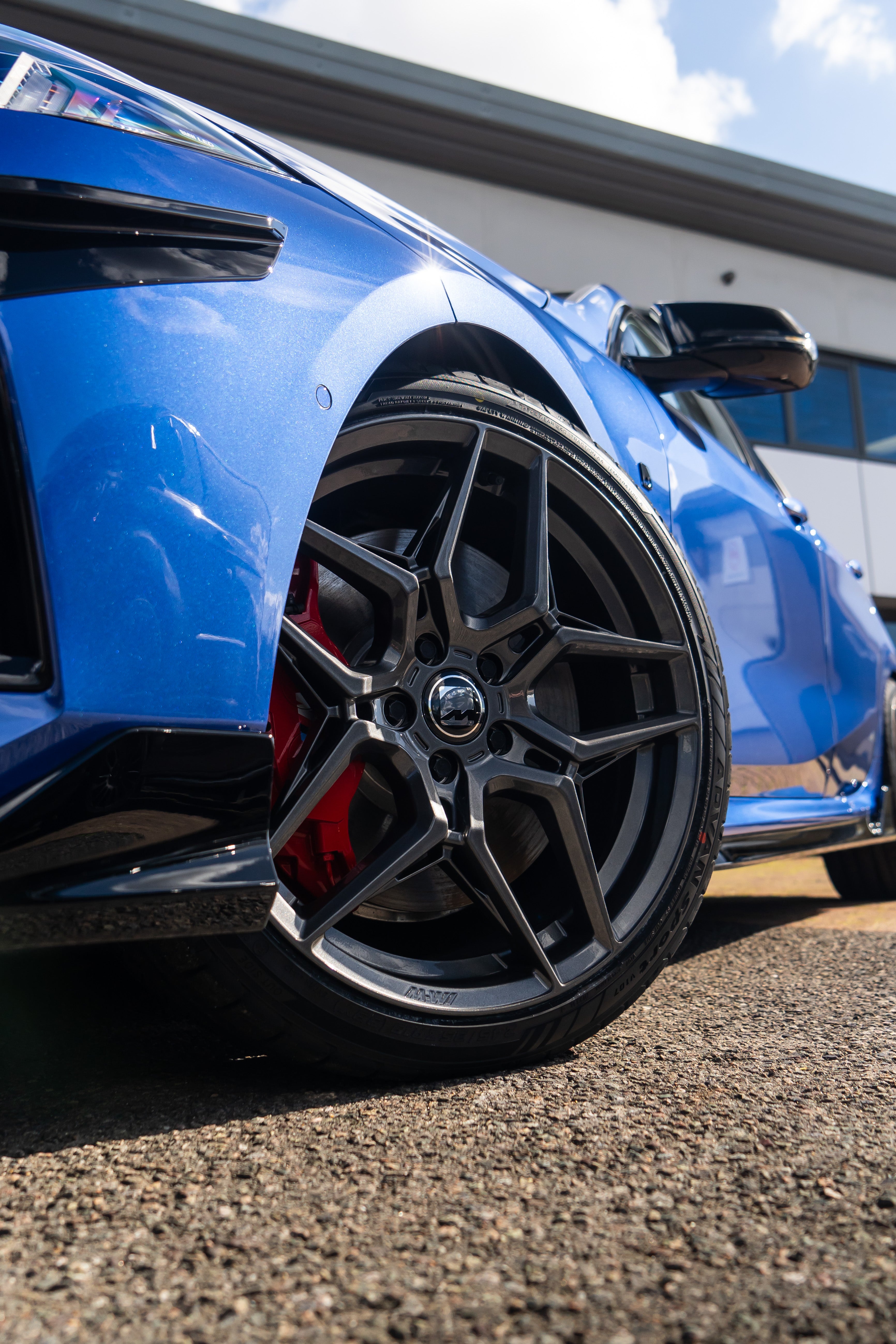 Carbon Grey M-W1 wheel on a blue BMW parked on a brick driveway.