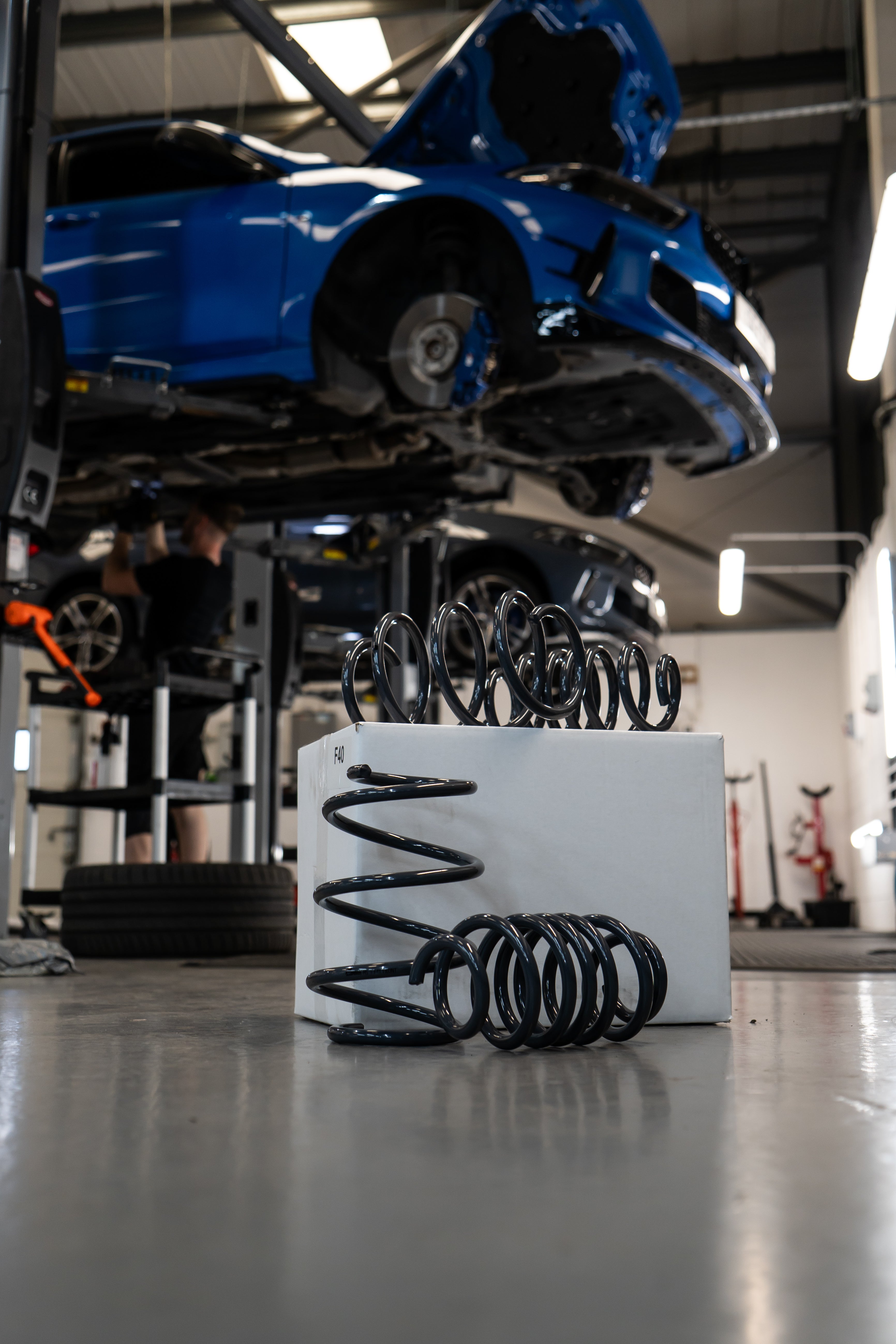 Lowering Springs for a car on a white box in the foreground, a car suspended in the air in a workshop in the background.