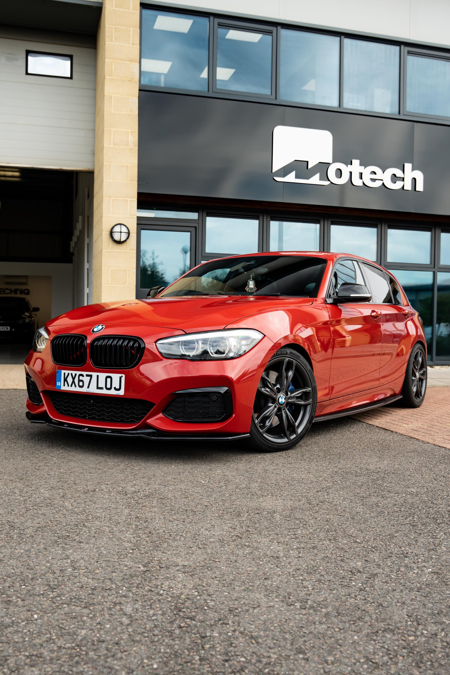 A red BMW 1 Series on Motech Stance + Springs parked outside a building with 'Motech' branding.