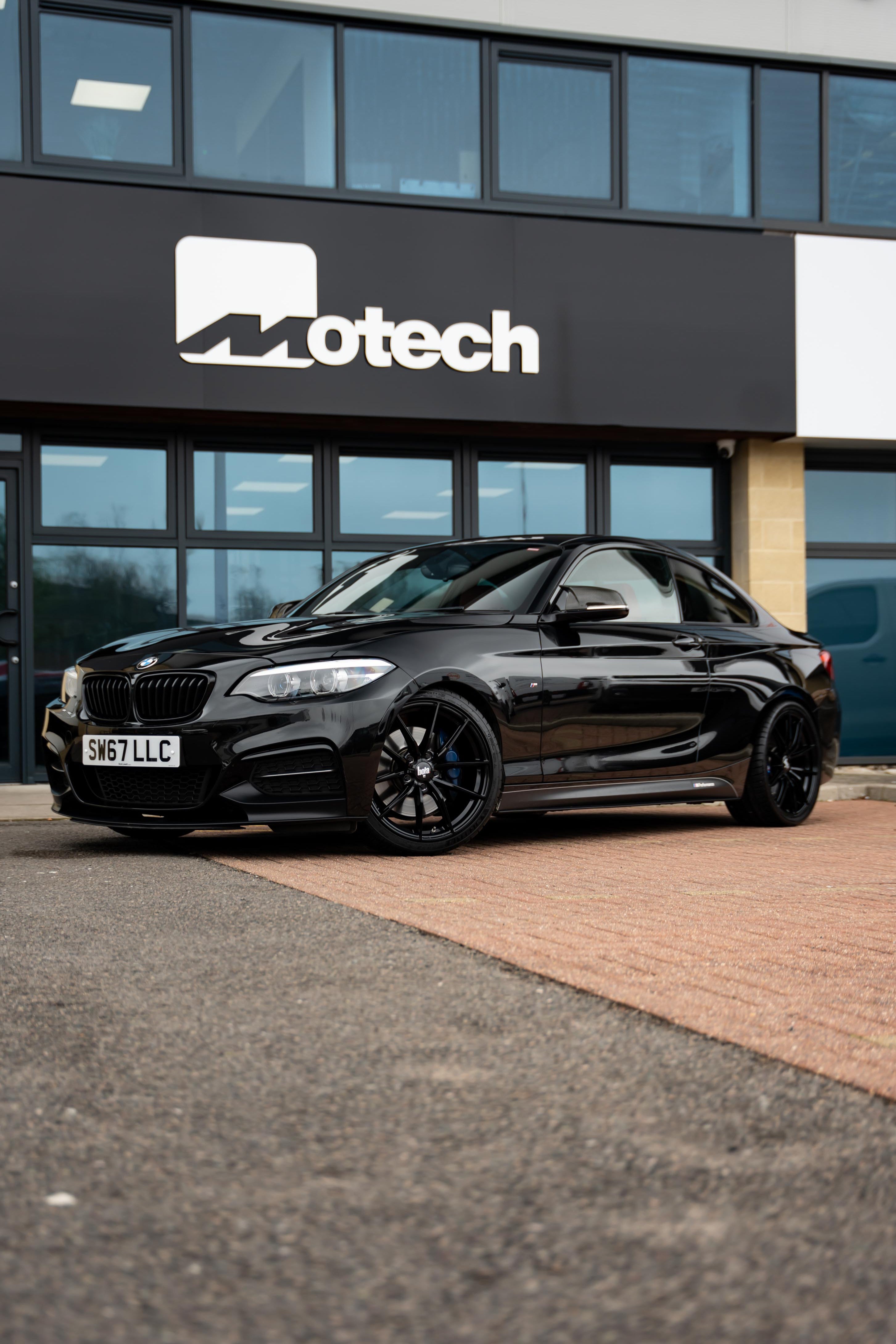 Black BMW car on Motech stance springs parked in front of a building with 'Motech' branding.