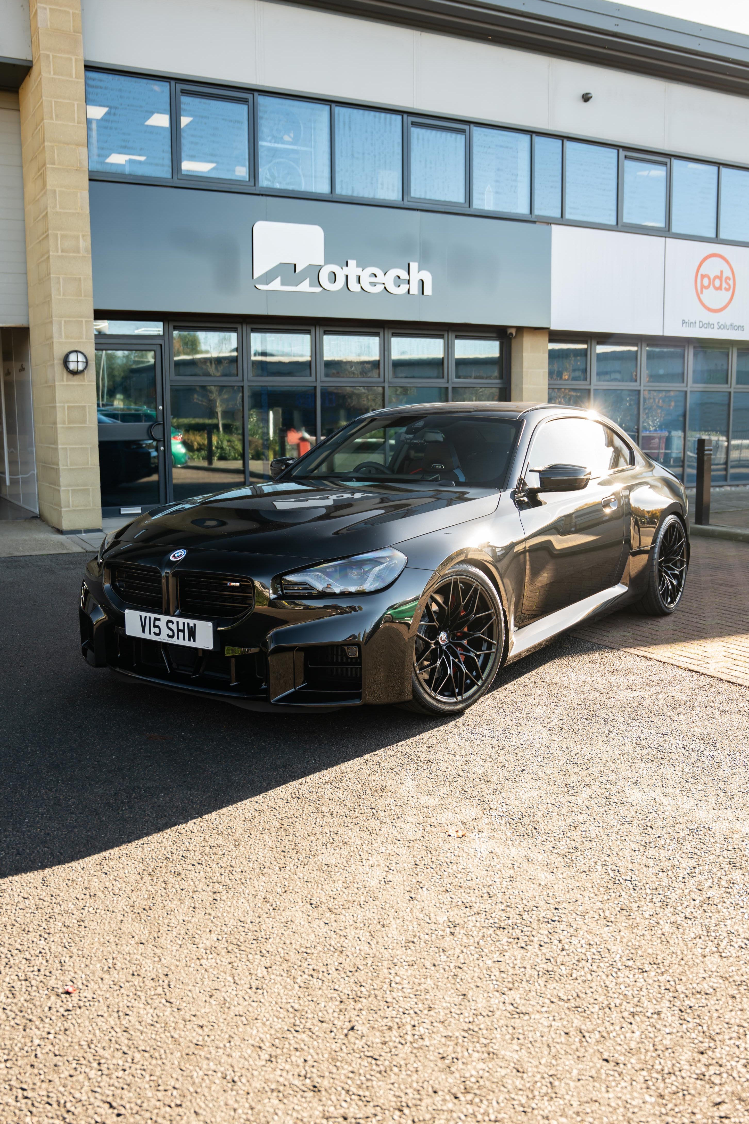 Black BMW M2 G87 on Motech Stance Springs parked outside a building with 'Motech' branding. 