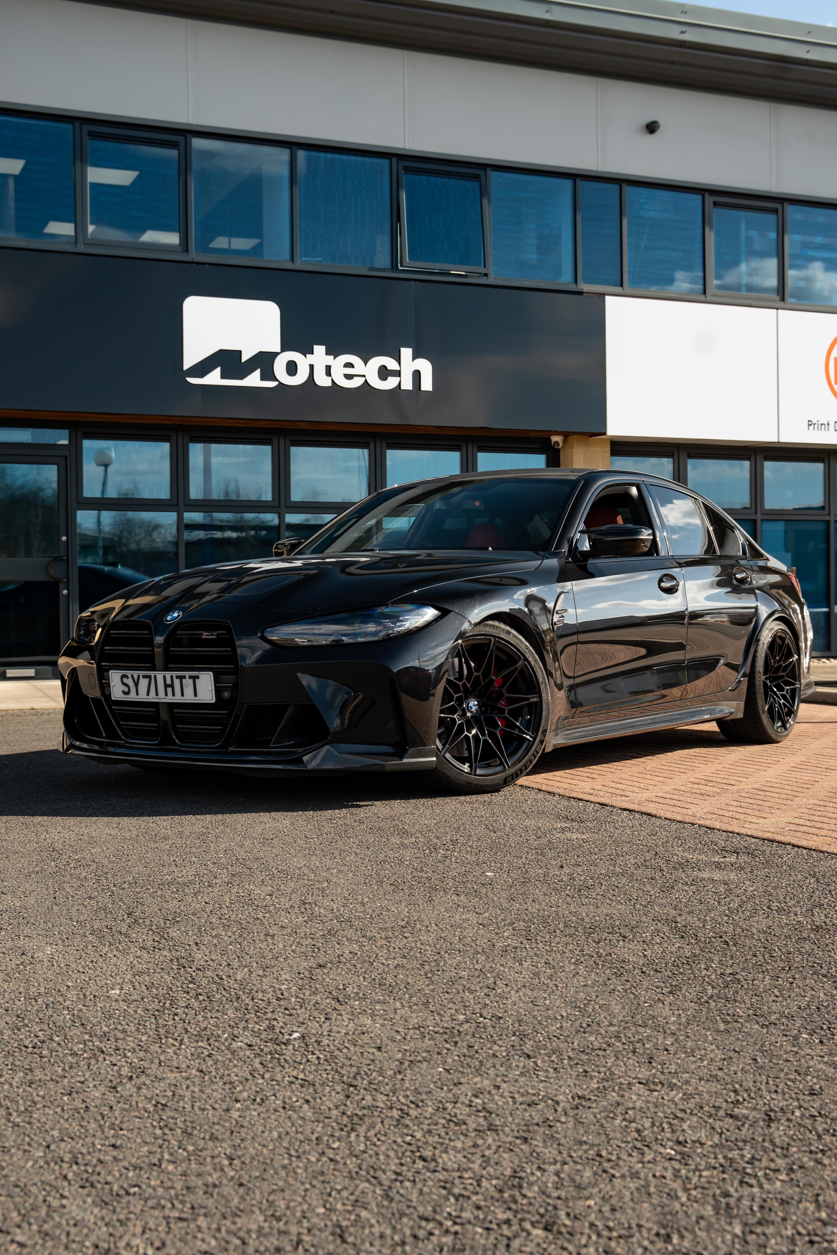 Black BMW M3 G80 on Motech Stance springs parked outside a building with 'Motech' branding. 