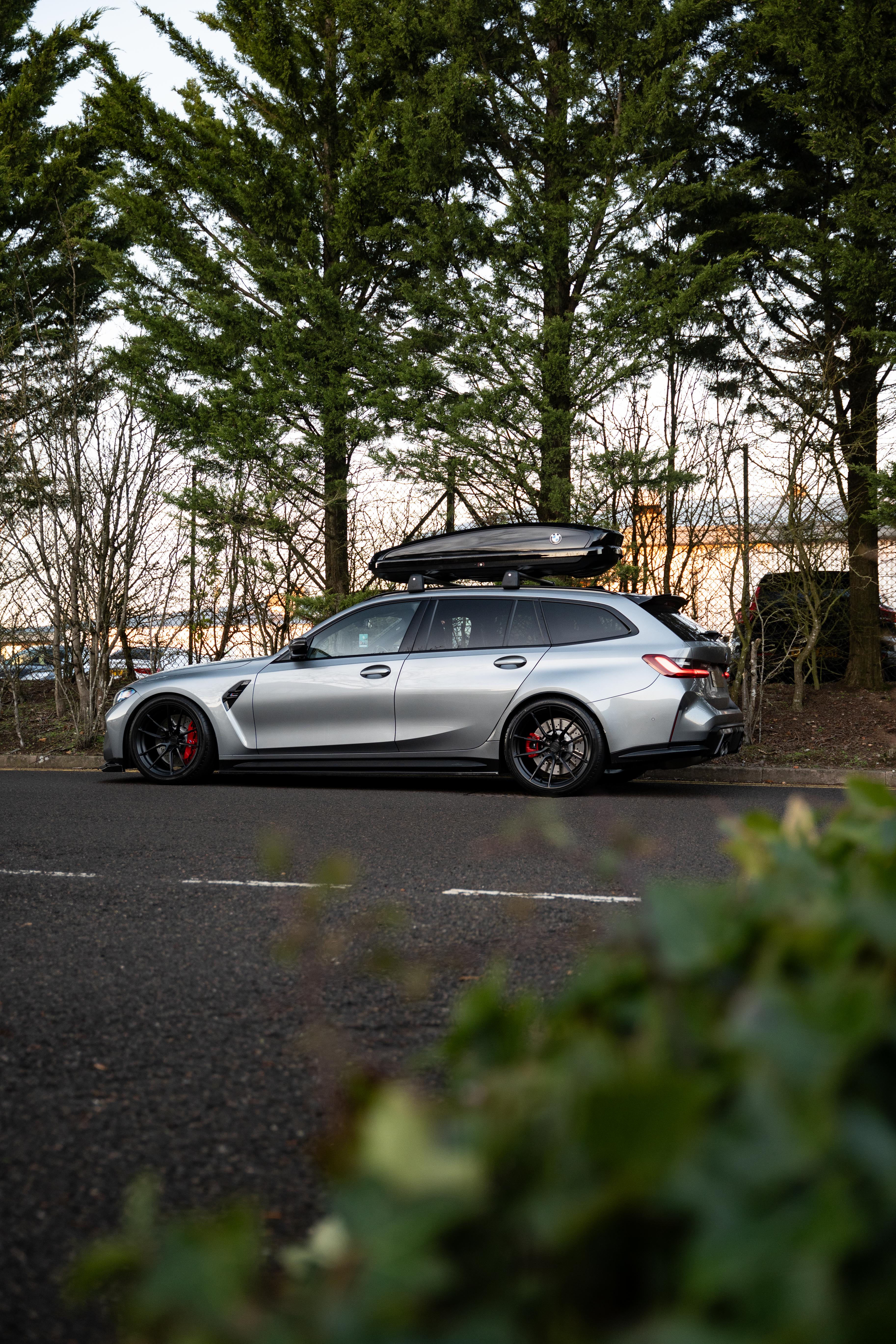 Side-on shot of a Grey G81 M3 Touring with a roof box & is sat on Motech stance springs - parked in front of some trees.