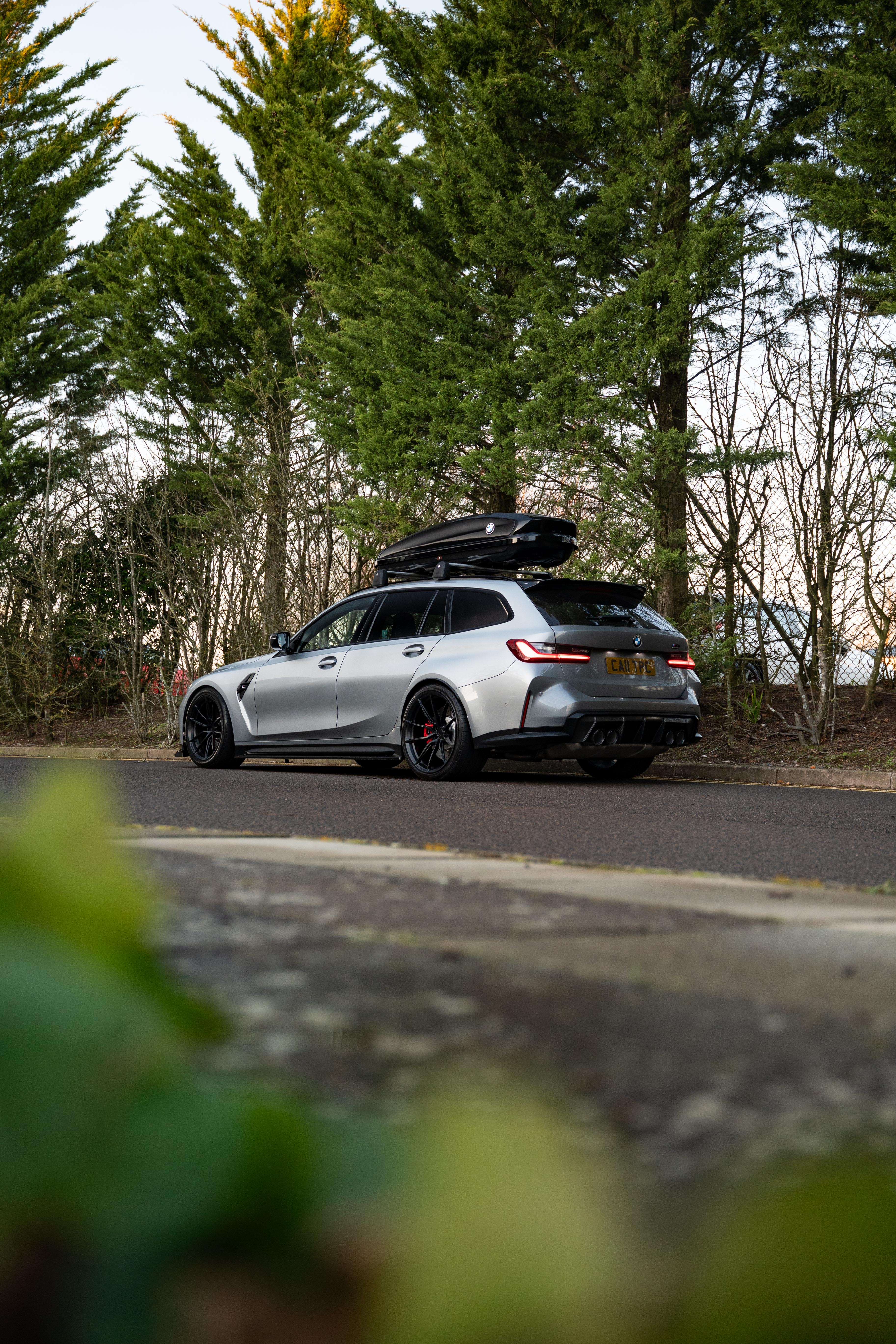 Grey G81 M3 Touring with a roof box & is sat on Motech stance springs - parked in front of some trees. 