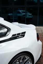 White car with a black decal on the side, parked in front of a glass building.