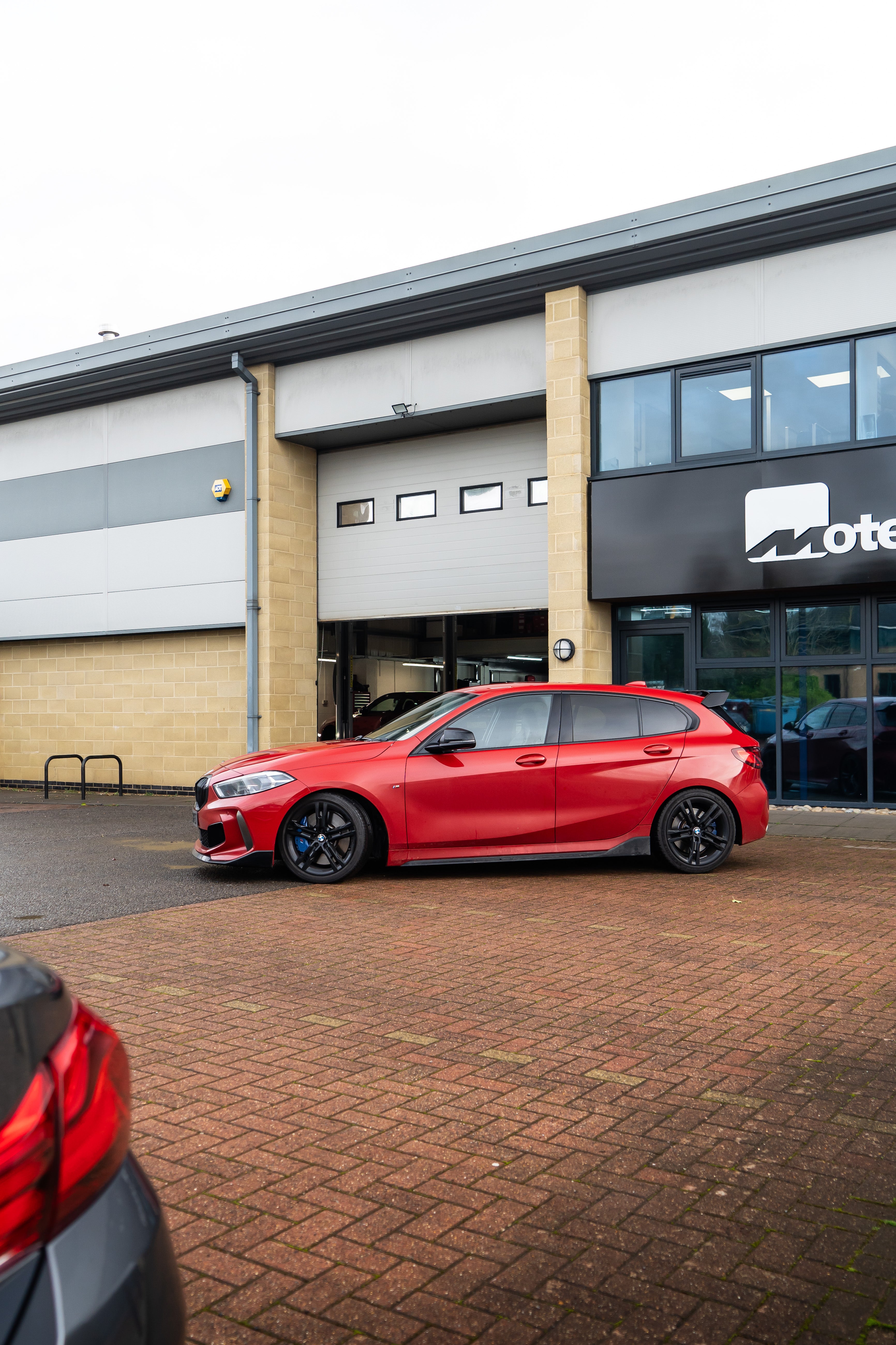 Side-on shot of a red BMW 1 Series on Motech Stance Springs with a carbon side skirt outside the 'Motech' building. 