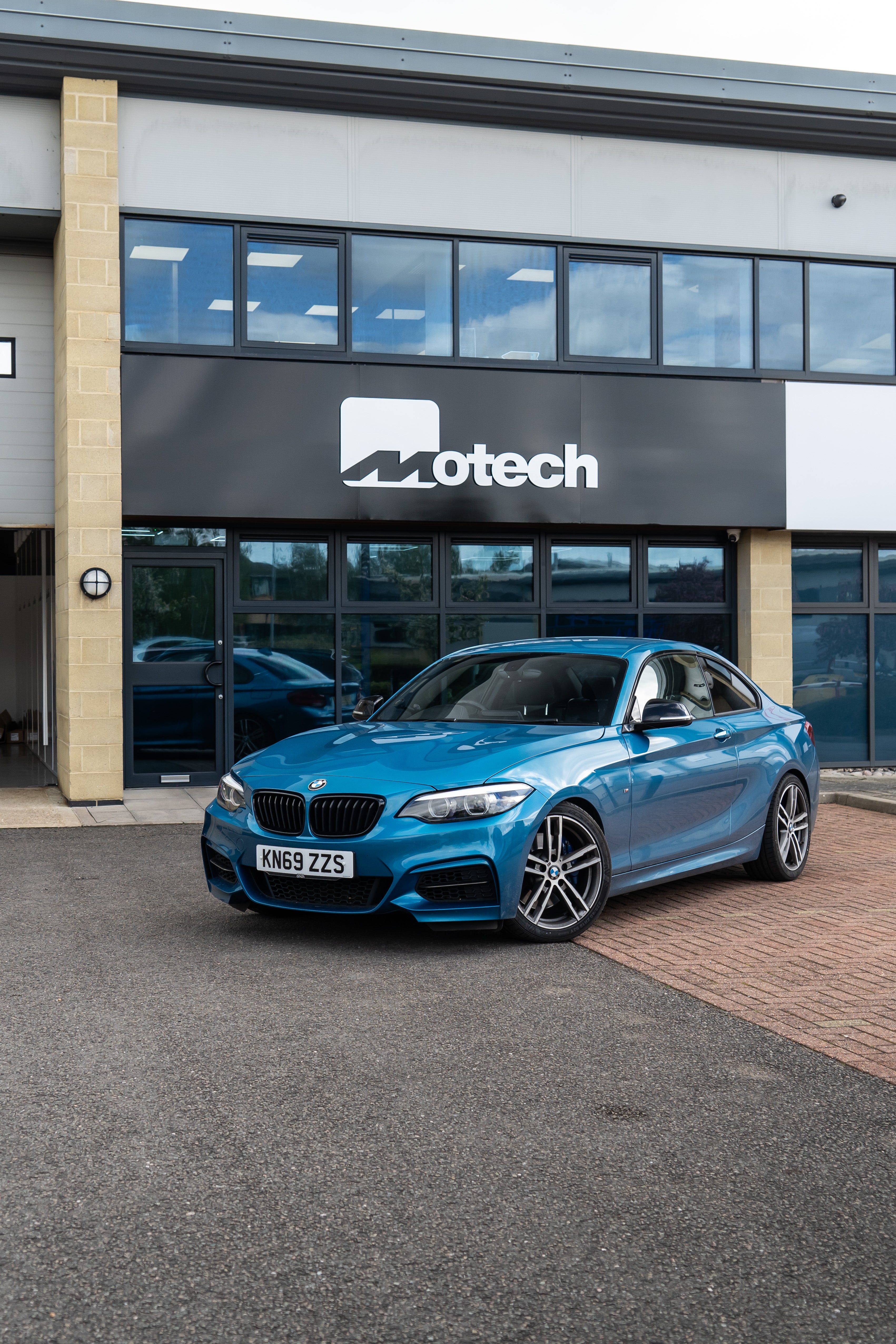 Blue BMW on Motech Stance Springs parked outside a building with 'Motech' branding. 