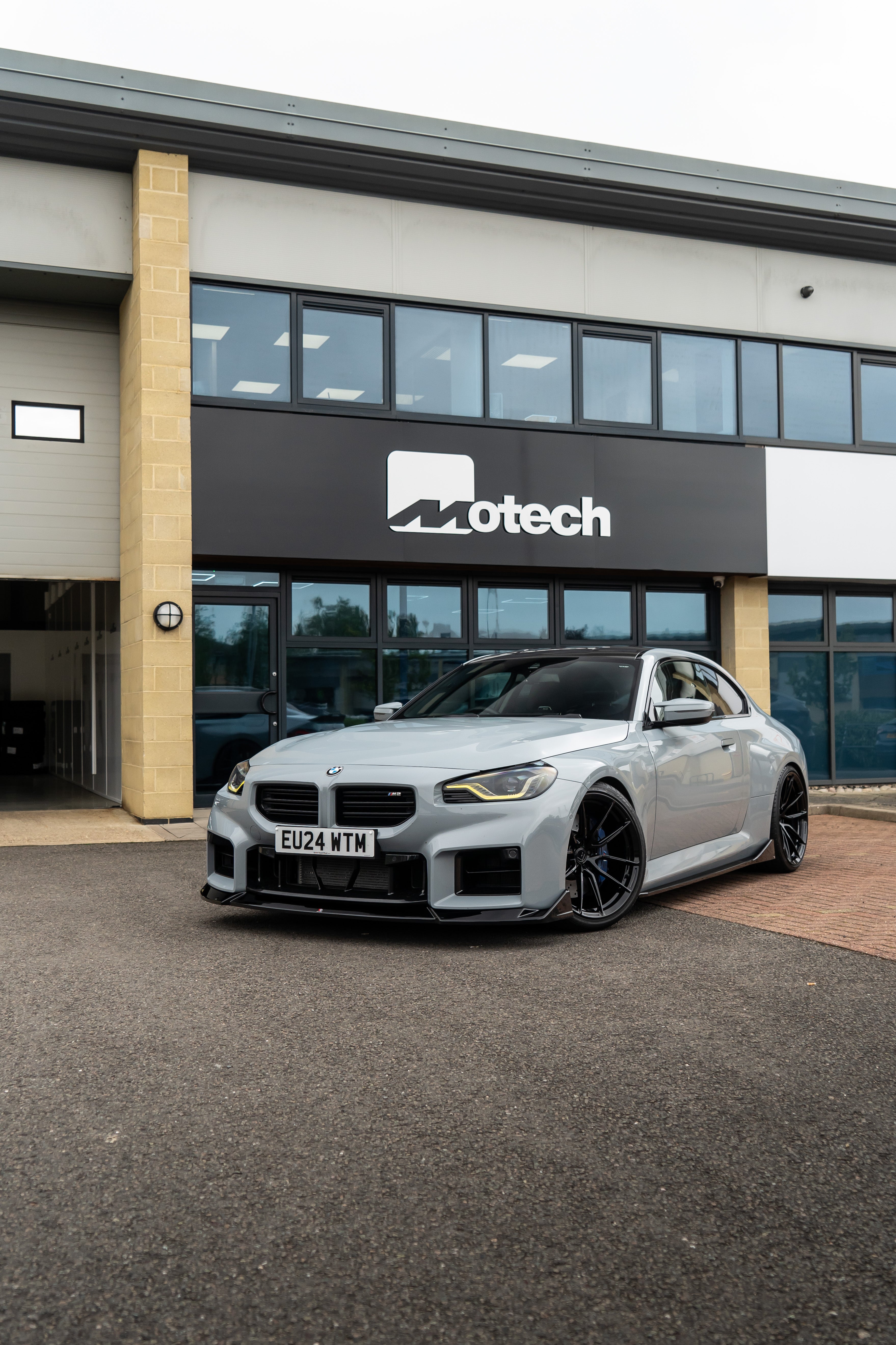 Grey BMW M2 G87 on Motech Stance Springs parked outside a building with 'Motech' branding. 