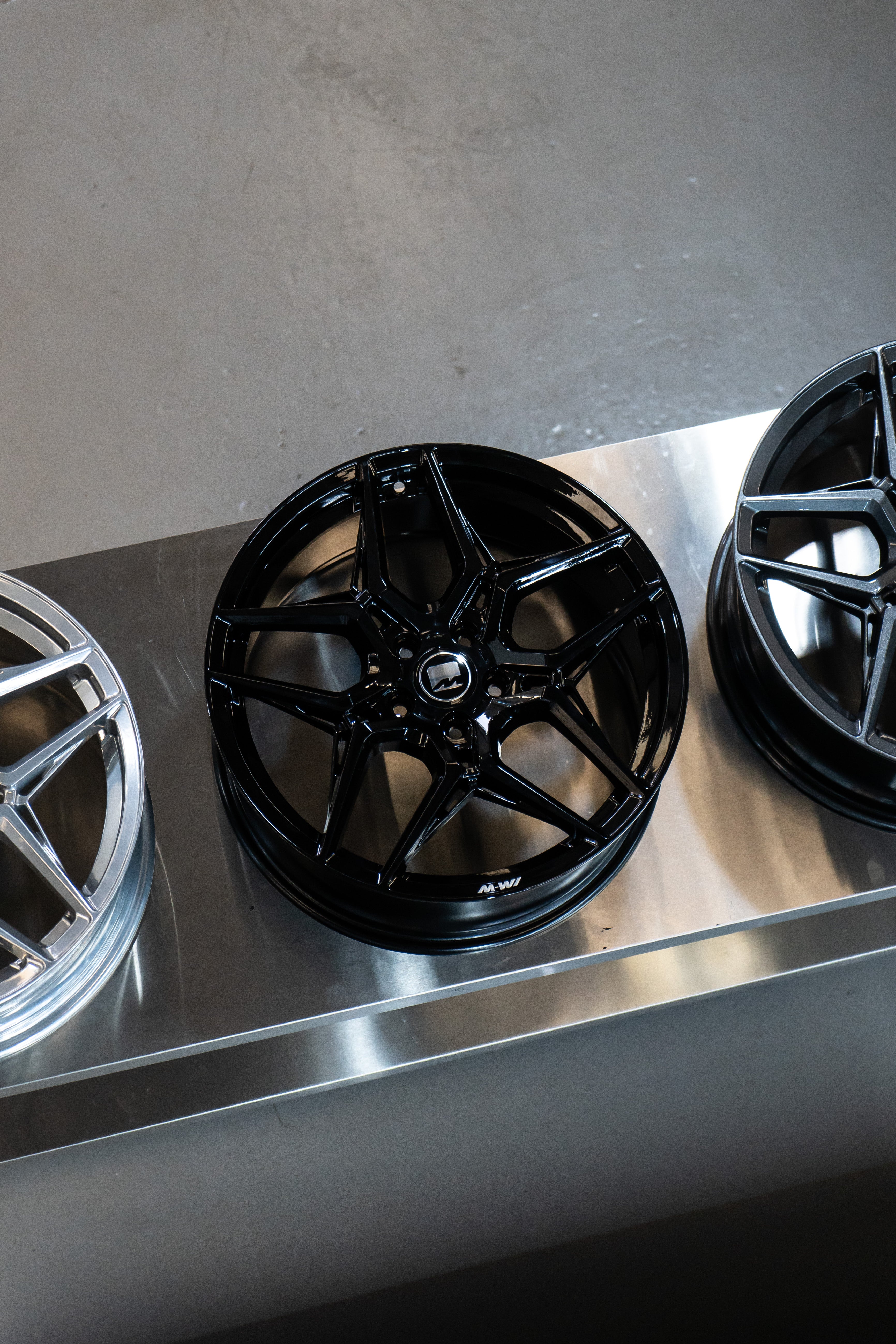 Precision Black M-W1 Wheel in a workshop beside 2 other wheels of different colour.