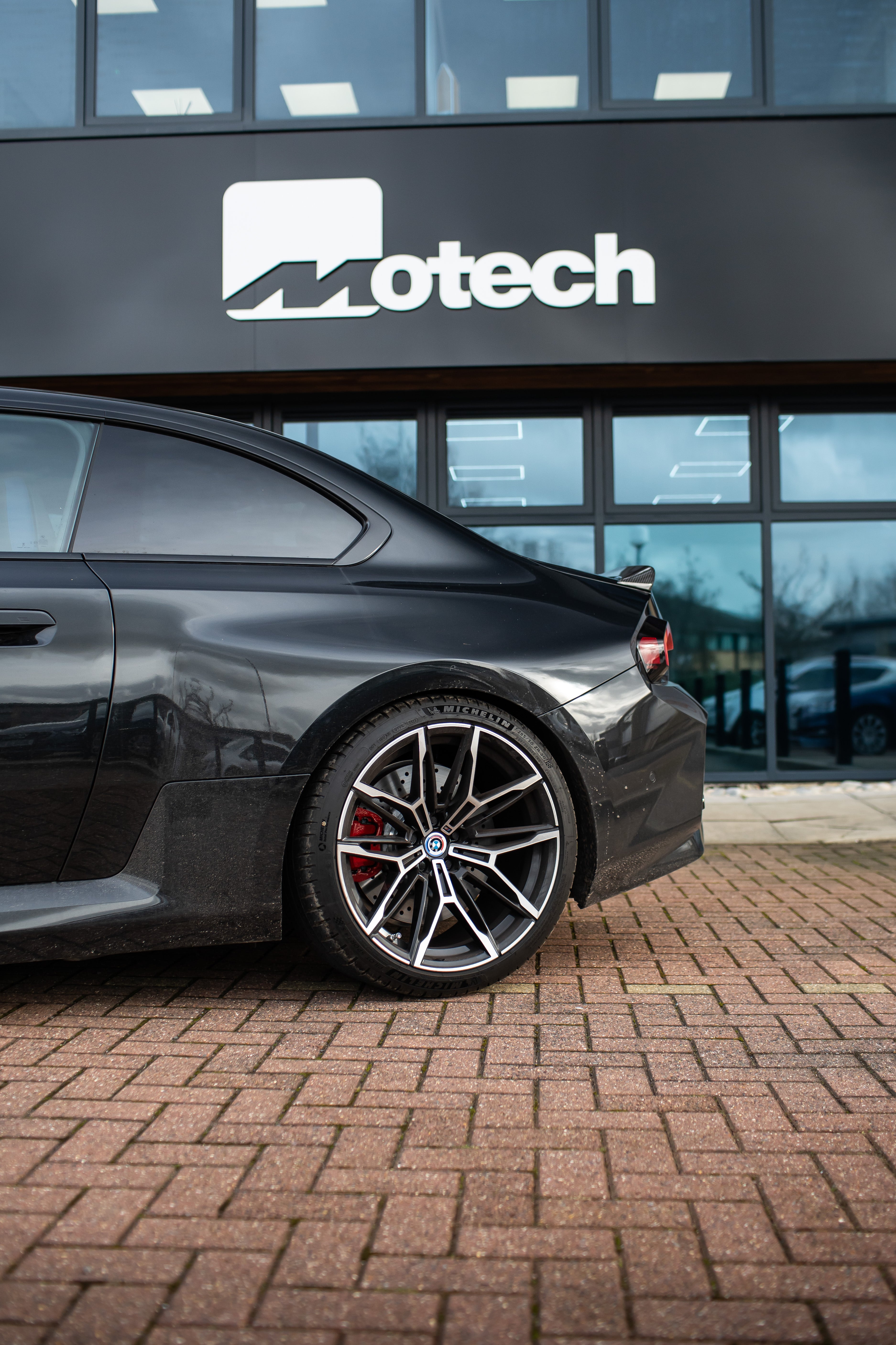 The back of a Black BMW M2 G87 on Motech Stance Springs parked outside a building with 'Motech' branding. 